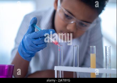 Un ragazzo dai capelli scuri in un camice da laboratorio che tiene una siringa con un reagente e che ha un aspetto occupato Foto Stock