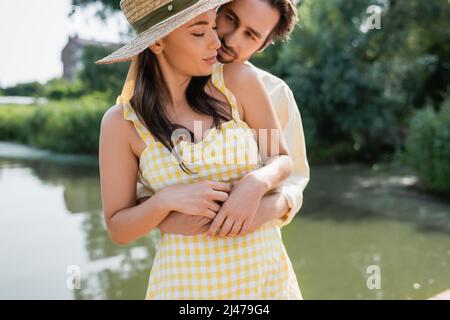 uomo bearded che abbraccia la ragazza in cappello di paglia vicino al lago Foto Stock