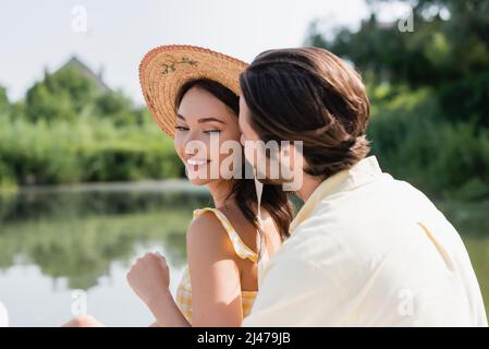 uomo sfocato sussurrando nell'orecchio di donna sorridente in cappello di paglia Foto Stock