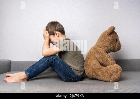 un bambino preschooler frustrato è seduto sul divano con la schiena a un grande orsacchiotto Foto Stock