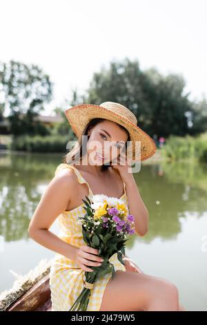 bella giovane donna in paglia cappello e vestito che tiene fiori mentre si siede vicino al lago Foto Stock