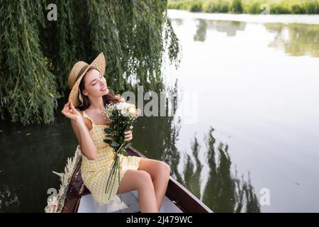 giovane donna sognante che regola il cappello di paglia e tiene i fiori durante il giro in barca sul lago Foto Stock