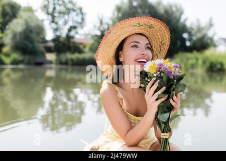 giovane donna gioiosa in cappello di paglia e vestito che tiene fiori vicino al lago Foto Stock