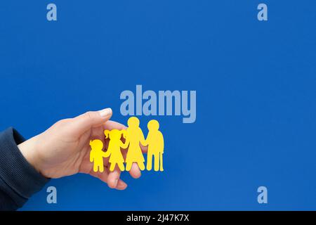 Mano che aiuta a tenere la famiglia Ucraina, silhouette di carta gialla. Piatto su sfondo blu. Aiuto volontario ai rifugiati ucraini. Foto Stock