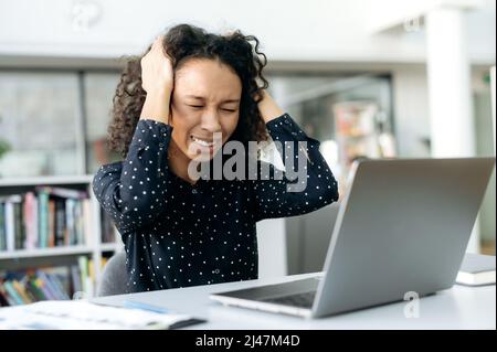 Overstressed, donna americana africana, direttore, seo, responsabile di accreditamento, seduta su una scrivania di fronte a un computer portatile, tenendo la testa, avendo un forte mal di testa, avendo bisogno di farmaci e facendo una pausa Foto Stock
