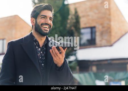 Un giovane uomo latino-americano bello in vestiti neri che tiene un telefono e sorridente ampiamente medio primo piano sfondo urbano esterno . Foto di alta qualità Foto Stock