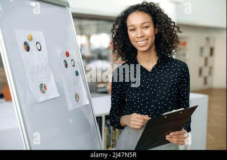 Smart auto-sicuro, ricci-capelli giovane adulto ragazza scura-skinned, collaboratore della società, responsabile delle vendite, si trova in ufficio vicino a una lavagna bianca, lavora con grafici e documenti, guarda la fotocamera, sorride Foto Stock