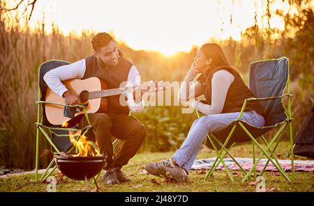 La vita è migliore intorno al fuoco del campeggio. Scatto di un tu che suona la sua chitarra a sua moglie fuori mentre campeggio. Foto Stock