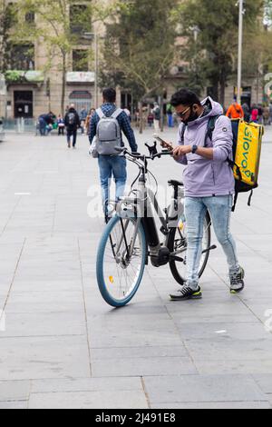 Corriere maschio in una maschera medica per consegnare il cibo. Deliveryman using smartphone.Barcelona, Spagna - 09 aprile 2022 Foto Stock