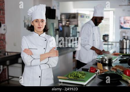 Ritratto di lavoratore di cucina ristorante sicuro in piedi con le braccia incrociate sorridendo alla macchina fotografica. Esperto di gastronomia che indossa uniforme da cucina mentre prepara erbe fresche per il servizio cena. Foto Stock