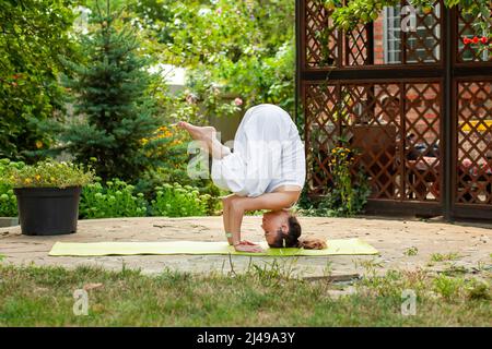 La giovane donna pratica lo yoga nel cortile di una casa di campagna. Supporto - Ardha Salamba Shirshasana Foto Stock