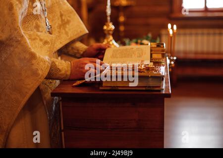 Foto ritagliata del prete in cassone cerimoniale d'oro stand al leggio con libri, lettura libro di preghiera in chiesa ortodossa. Foto Stock