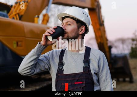 Costruttore in casco bere caffè da tazza di carta in pausa caffè. Escavatore sullo sfondo. Foto Stock