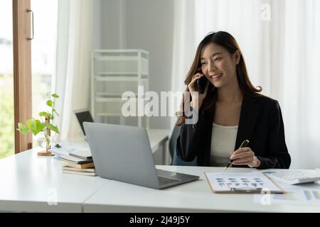 Asian Businesswoman che parla sul telefono mobile che lavora sul laptop per la finanza in ufficio moderno. Foto Stock