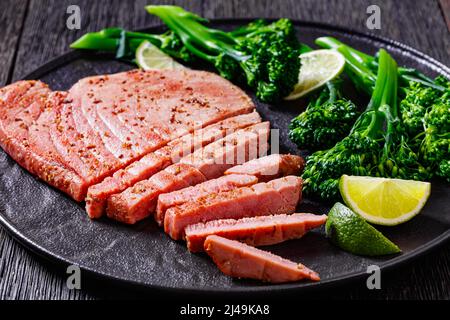 bistecca di tonno fritta con broccolini parboiled e lime su piatto nero su tavola di legno scuro, vista orizzontale dall'alto Foto Stock