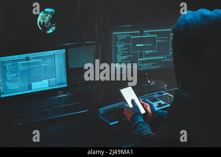 Hacker di testa in Hood lavorando al computer portatile e cellulare digitando testo in stanza buia, un hacker anonimo utilizza malware con il telefono cellulare per hack Foto Stock