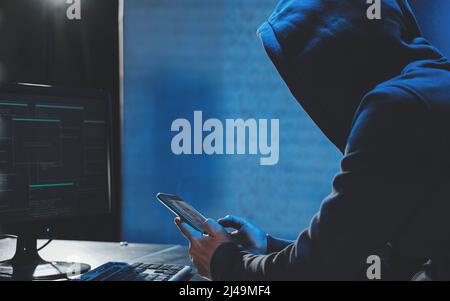 Hacker di testa in Hood lavorando al computer e telefono cellulare digitando testo in camera oscura, un hacker anonimo utilizza malware con il telefono cellulare per hack Foto Stock