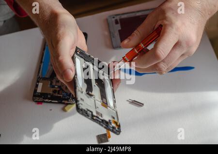 Repair of smartphones concept. A man unscrews the screws fixing the display from the board. Part of a series. Inside the room. Selective focus. Foto Stock