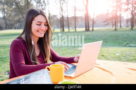 Una giovane bella donna lavora a distanza in un parco in una giornata di sole. Utilizza un computer portatile e auricolari bianchi. Ci sono delle tazze sul tavolo. Lavoro in remoto Foto Stock