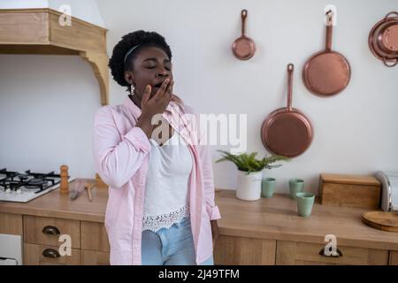 Una donna con pelle scura in piedi in cucina e sbadiglio Foto Stock