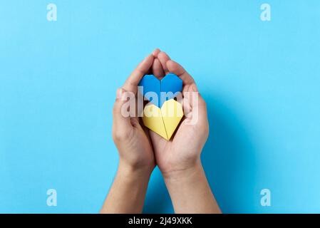 Mani di una persona sconosciuta che tiene e protegge un paio di cuori di carta blu e giallo, colori della bandiera Ucraina. Immagine simbolica a supporto di U Foto Stock