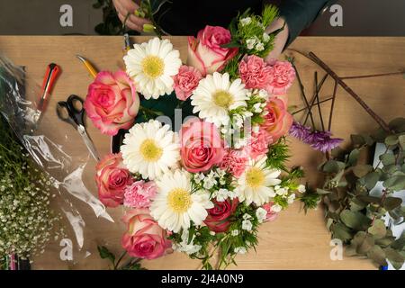 Vista dall'alto delle mani di una giovane fiorista femminile che crea una bella composizione di delicate rose rosa e margherite bianche sul tavolo Foto Stock