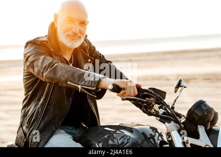 Uomo anziano audace che indossa una giacca in pelle che guida la motocicletta all'aperto il giorno d'estate Foto Stock