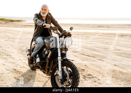 Uomo anziano audace che indossa una giacca in pelle che guida la motocicletta all'aperto il giorno d'estate Foto Stock