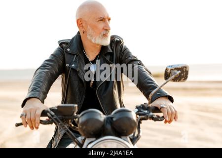 Uomo anziano audace che indossa una giacca in pelle che guida la motocicletta all'aperto il giorno d'estate Foto Stock