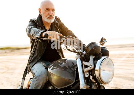 Uomo anziano audace che indossa una giacca in pelle che guida la motocicletta all'aperto il giorno d'estate Foto Stock