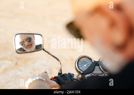 Uomo anziano audace che guarda lo specchietto retrovisore mentre guida la moto la giornata estiva Foto Stock