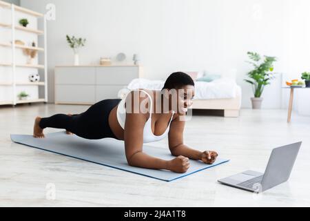 Sport online e perdita di peso. Sovrappeso giovane donna nera che fa esercizi di fronte al portatile a casa, a tutta lunghezza Foto Stock