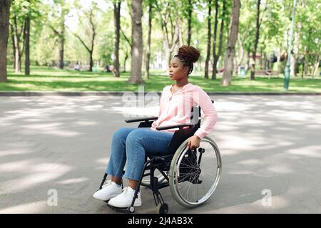Intera lunghezza di giovane donna nera in sedia a rotelle andando a piedi al parco urbano, spazio vuoto Foto Stock