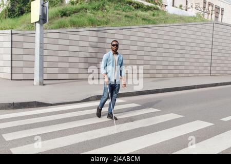 Giovane uomo nero con disabilità visiva indossando occhiali scuri, usando bastone da passeggio per attraversare la strada nel centro della città Foto Stock