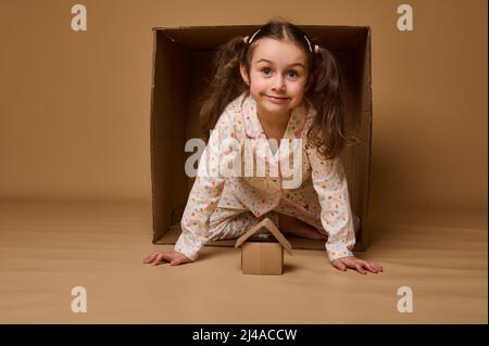 Bella bambina che sbuccia fuori da dietro una scatola di cartone, guardando la macchina fotografica. Scatola di cartone artigianale su sfondo beige con spazio per la copia degli annunci. Housi Foto Stock