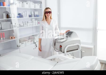 Donna gioiosa beautician in piedi vicino a macchina di epilazione laser Foto Stock