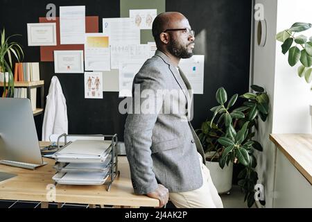 Medico generale africano appoggiato sul suo ufficio e guardando la finestra con vista pensiva Foto Stock