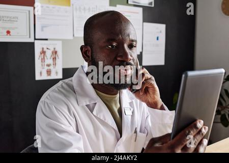 Medico africano di sesso maschile in cappotto bianco seduto sul posto di lavoro e tenendo il tablet pc davanti al suo volto mentre si ha una conversazione sul telefono cellulare Foto Stock