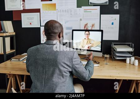 Vista posteriore dello specialista medico africano che parla con il suo paziente online sul monitor del computer mentre è seduto alla scrivania dell'ufficio Foto Stock