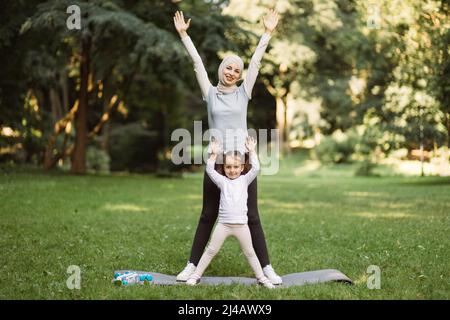 Attività fitness all'aperto. Simpatica donna musulmana in abbigliamento sportivo e la sua bambina prescolare che fa esercizi sportivi, in piedi con le braccia sollevate insieme nel parco estivo Foto Stock