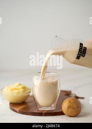 Latte di patate versato in vetro su sfondo di marmo bianco. Versare il latte vegano in vetro, con purea di patate e tuberi di patate sullo sfondo. Spazio di copia. Foto Stock