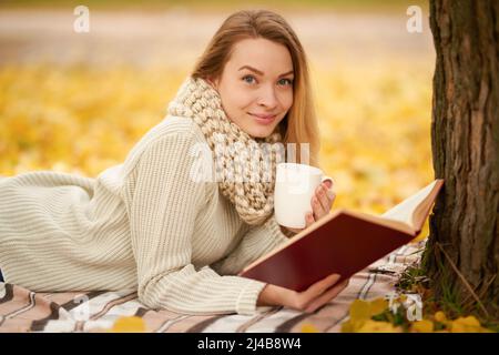 Una bella ragazza legge un libro e beve tè caldo, shikolad caldo, caffè in un caldo parco autunnale. Concetto di calore, atmosfera e comfort autunnali. Con Foto Stock