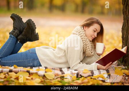 Una bella ragazza legge un libro e beve tè caldo, shikolad caldo, caffè in un caldo parco autunnale. Concetto di calore, atmosfera e comfort autunnali. Con Foto Stock