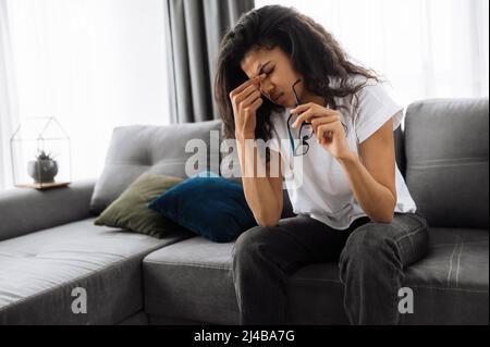 Studentessa, lavoratore o casalinga esausta tiene gli occhiali in mano, seduto sul divano a casa, stanca da lavoro a distanza, studio o lavori domestici, massaggia gli occhi, ha bisogno di riposo, lavoro in eccesso Foto Stock