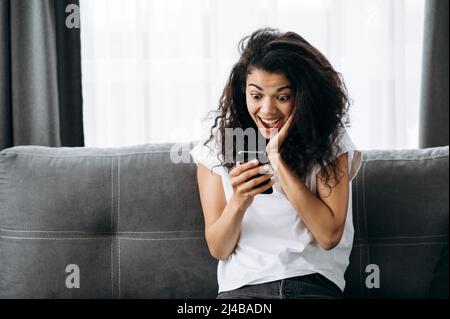 Ritratto di bella giovane afroamericana a casa. La ragazza si siede sul divano e usa il suo telefono. Donna gioiosa guardare lo schermo, leggere buone notizie, fa un buon acquisto online o ottenere un Foto Stock