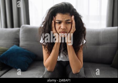 Primo piano di una triste giovane afroamericana. Brunette annoiato a casa, stanco o stressato. Ragazza turbata seduta sul divano, avendo un mal di testa o sensazione di depressione Foto Stock