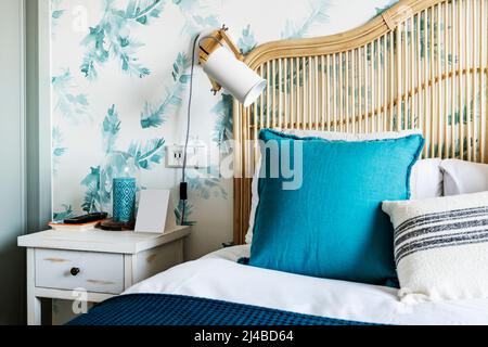 Dettaglio della testiera in bambù e vimini in una camera da letto con cuscini blu e bianchi, un comodino in legno e carta da parati sulla parete Foto Stock