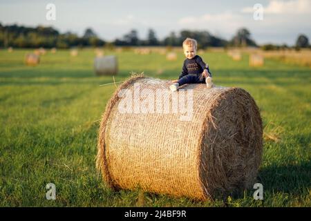 Il ragazzino siede su un grande rotolo di fieno in un prato verde, posando splendidamente con un sorriso. Foto Stock