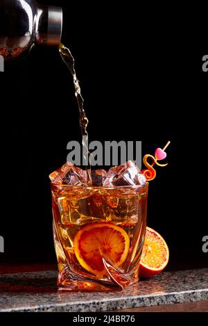 Cocktail di alcol che si versa dallo shaker nel gless con ghiaccio a buccia d'arancia su un tavolo di pietra. Foto Stock