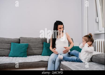 Una donna caucasica incinta e di buona qualità tocca il suo grande pancino con entrambe le mani, sorride e guarda la figlia che le copre le orecchie con le mani. Foto Stock